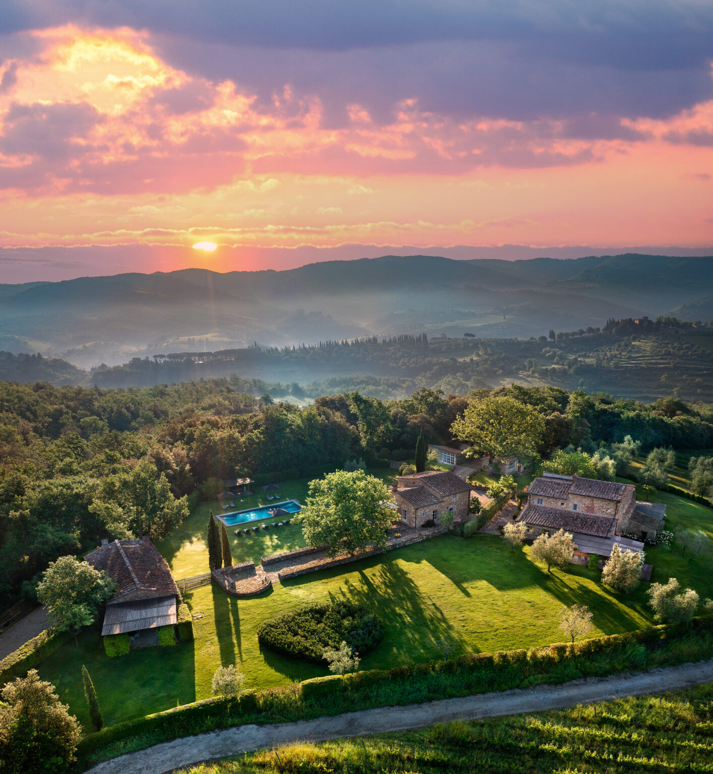 Aerial view of a picturesque countryside villa in Tuscany at sunset, featuring lush green gardens, a swimming pool, and rustic stone buildings surrounded by rolling hills and trees. The vibrant sky showcases hues of pink and orange, creating a serene and tranquil atmosphere. | Only on SopranoVillas