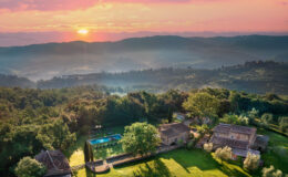 Aerial view of a picturesque countryside villa in Tuscany at sunset, featuring lush green gardens, a swimming pool, and rustic stone buildings surrounded by rolling hills and trees. The vibrant sky showcases hues of pink and orange, creating a serene and tranquil atmosphere.