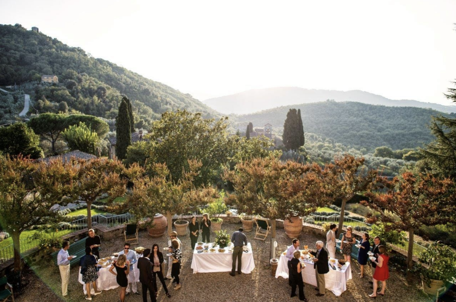 Outdoor wedding reception set in a scenic landscape, featuring guests mingling and enjoying food and drinks, surrounded by lush greenery and mountains under a soft sunset. | Only on SopranoVillas