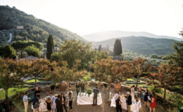 Outdoor wedding reception set in a scenic landscape, featuring guests mingling and enjoying food and drinks, surrounded by lush greenery and mountains under a soft sunset.
