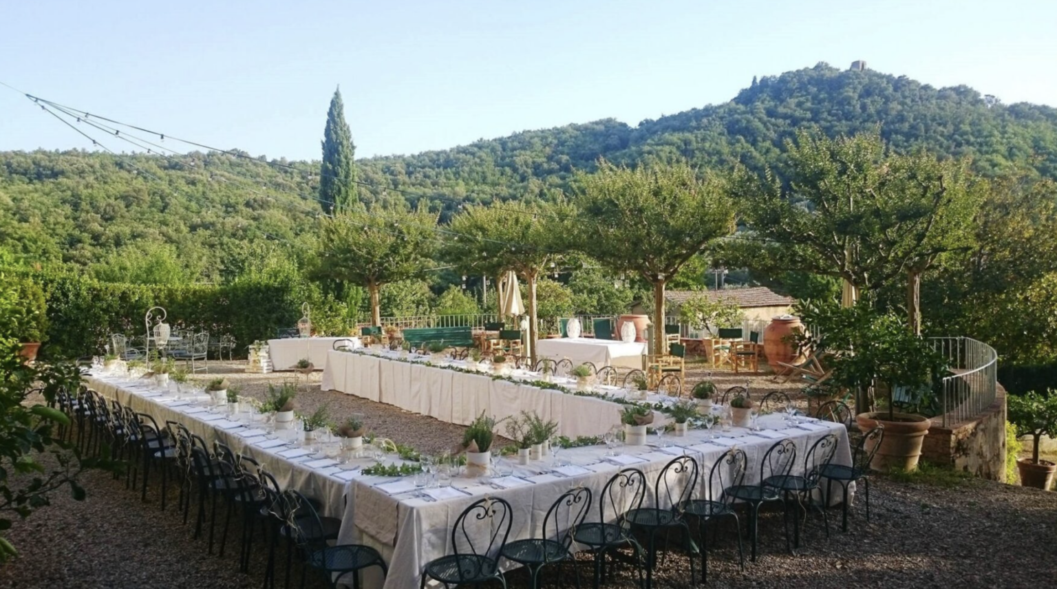 Outdoor dining setup with a long table elegantly arranged for an event, surrounded by lush greenery and hills, creating a picturesque setting. | Only on SopranoVillas