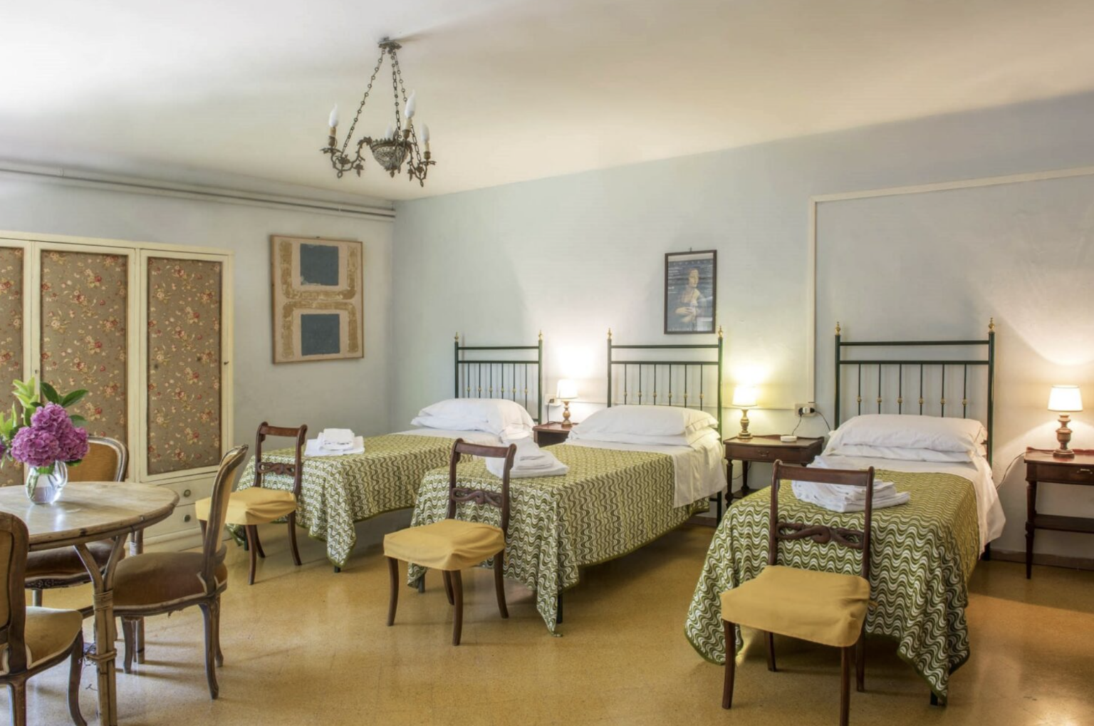 Cozy hotel room featuring two twin beds with patterned bedspreads, a wooden table and chairs, and soft lighting. Ideal for families or groups seeking comfortable accommodations. | Only on SopranoVillas