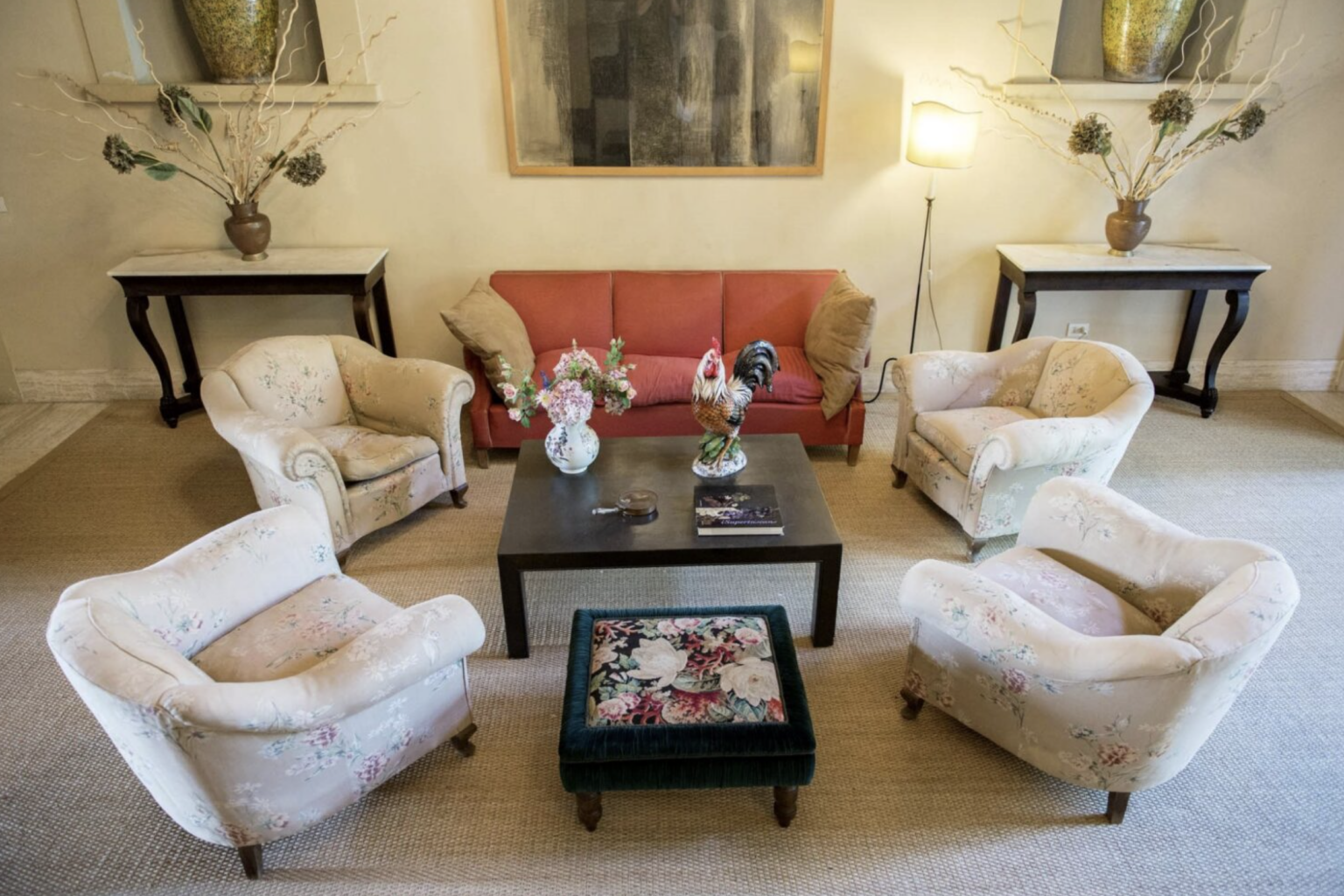 Cozy living room setup featuring a red sofa and floral armchairs arranged around a dark coffee table. A decorative rooster figurine and a vase of flowers are on the table, complemented by a plush ottoman with floral upholstery. Natural light illuminates the space, enhancing the inviting atmosphere. | Only on SopranoVillas