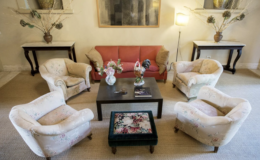 Cozy living room setup featuring a red sofa and floral armchairs arranged around a dark coffee table. A decorative rooster figurine and a vase of flowers are on the table, complemented by a plush ottoman with floral upholstery. Natural light illuminates the space, enhancing the inviting atmosphere.