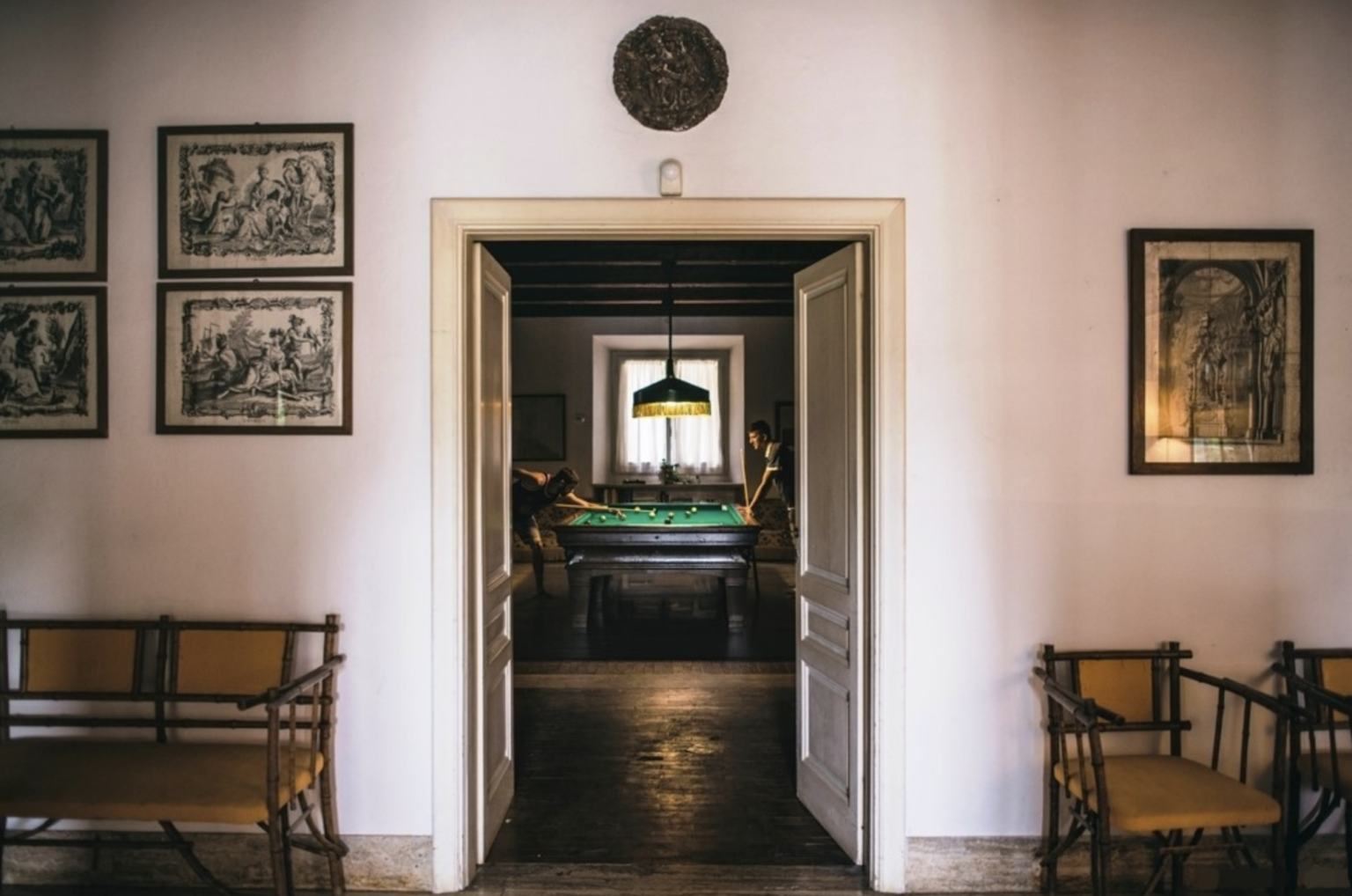 Interior view of a stylish room featuring a doorway leading to a billiards table area, with two individuals playing pool. The walls are adorned with framed artwork, and there are wooden benches with yellow cushions in the foreground, creating a cozy and inviting atmosphere. | Only on SopranoVillas