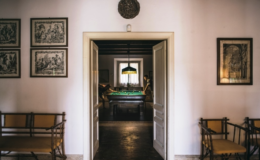 Interior view of a stylish room featuring a doorway leading to a billiards table area, with two individuals playing pool. The walls are adorned with framed artwork, and there are wooden benches with yellow cushions in the foreground, creating a cozy and inviting atmosphere.