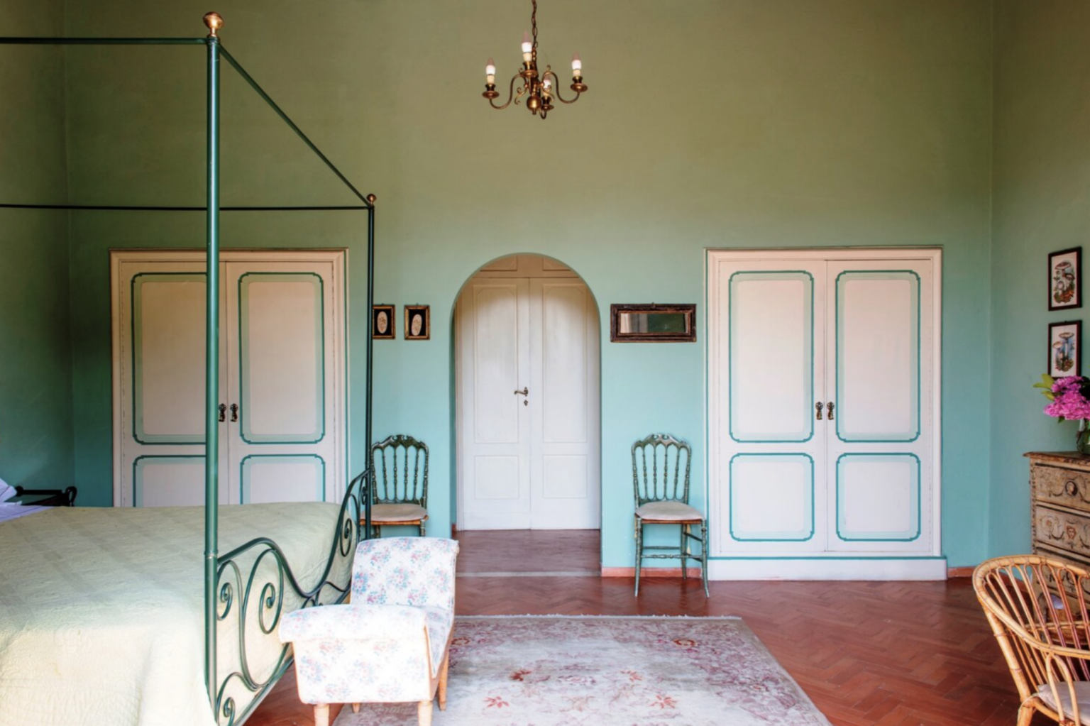 Elegant bedroom interior featuring a pastel green color scheme, a four-poster bed, wooden furniture, and decorative wall art. The room includes two white doors, vintage chairs, and a chandelier, creating a cozy and inviting atmosphere. | Only on SopranoVillas