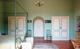 Elegant bedroom interior featuring a pastel green color scheme, a four-poster bed, wooden furniture, and decorative wall art. The room includes two white doors, vintage chairs, and a chandelier, creating a cozy and inviting atmosphere.