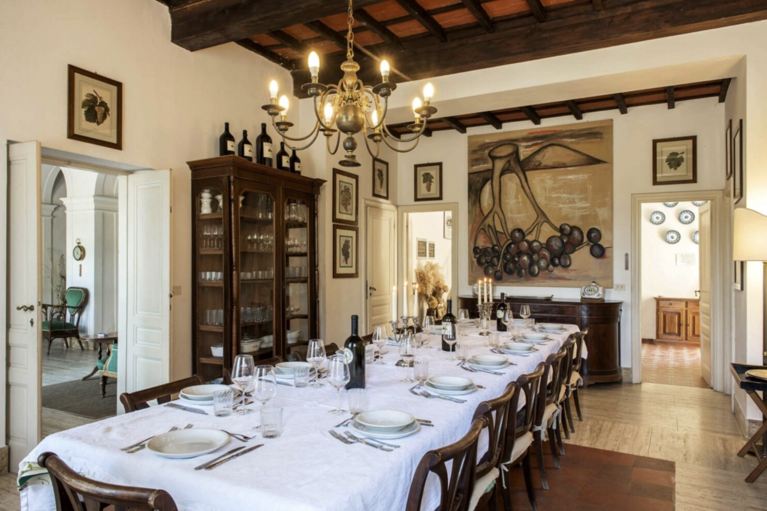 Elegant dining room featuring a long table set for a meal, adorned with white tablecloth, fine china, and glassware. The room showcases rustic wooden beams, vintage artwork on the walls, and a chandelier, creating a warm and inviting atmosphere. | Only on SopranoVillas