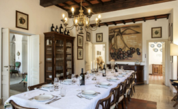 Elegant dining room featuring a long table set for a meal, adorned with white tablecloth, fine china, and glassware. The room showcases rustic wooden beams, vintage artwork on the walls, and a chandelier, creating a warm and inviting atmosphere.