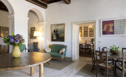 Cozy interior of a stylish living room featuring a wooden dining table, green sofa, and decorative artwork on the walls, with a vase of flowers as the centerpiece. The room is well-lit, showcasing an inviting atmosphere with a glimpse of another dining area through an open doorway.