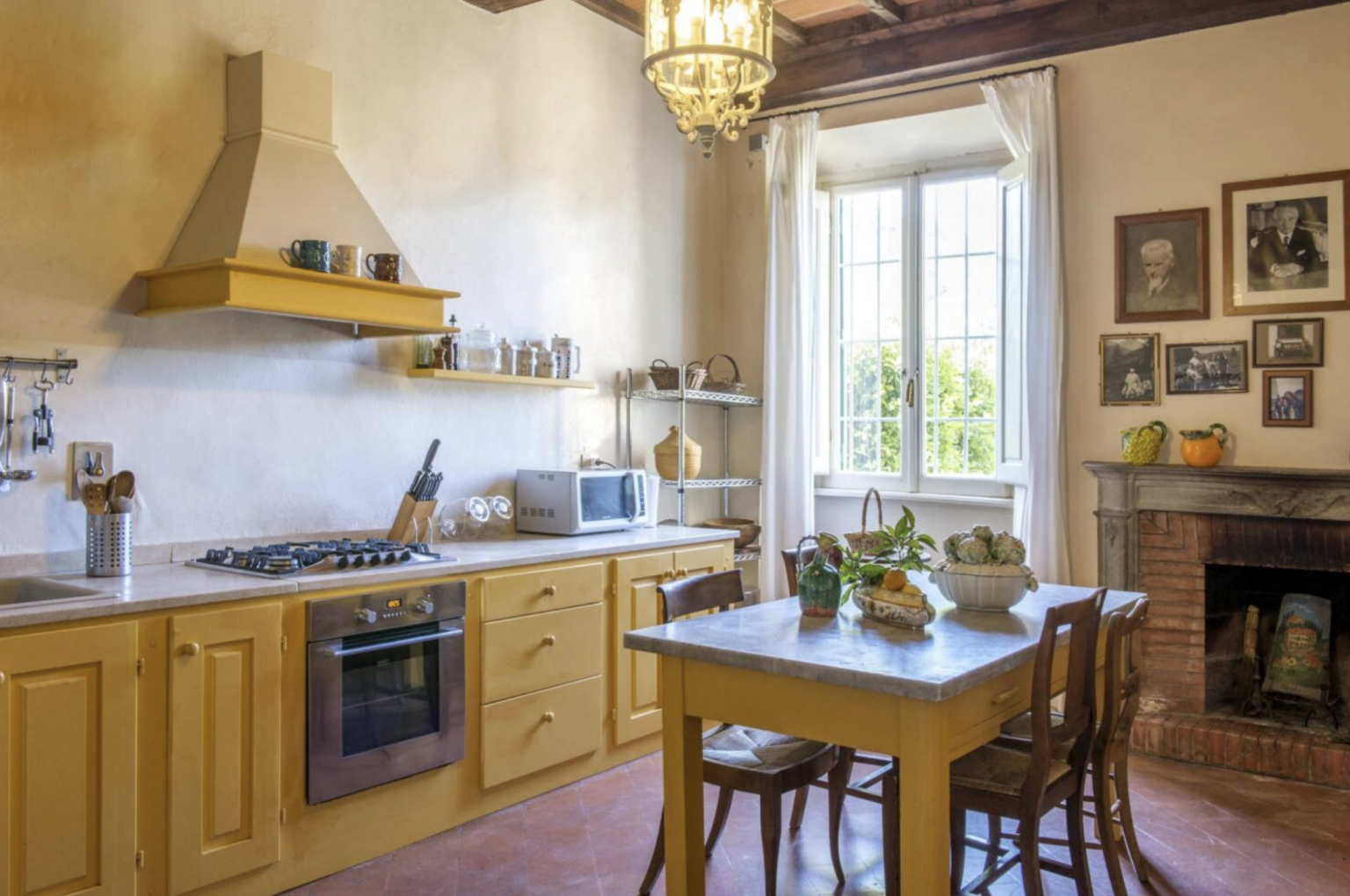 Bright and inviting kitchen featuring yellow cabinetry, a gas stove, and a marble-topped dining table. Natural light streams through large windows, illuminating the rustic decor, including framed photographs and a classic fireplace. | Only on SopranoVillas