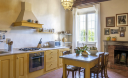 Bright and inviting kitchen featuring yellow cabinetry, a gas stove, and a marble-topped dining table. Natural light streams through large windows, illuminating the rustic decor, including framed photographs and a classic fireplace.