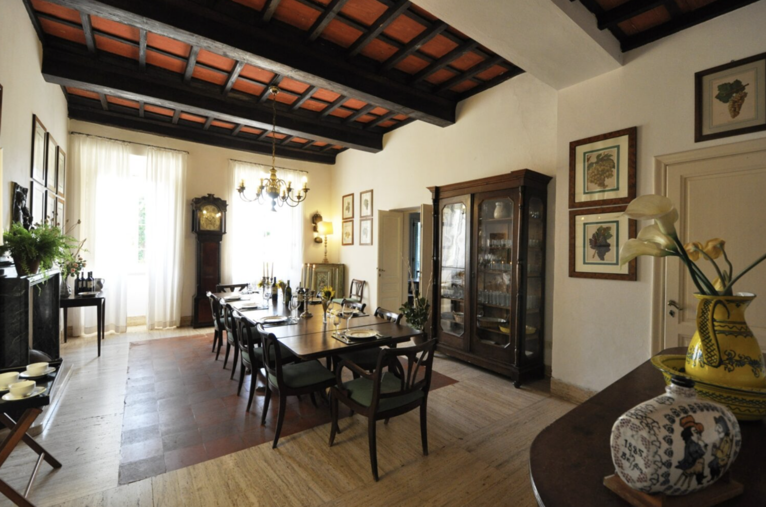 Elegant dining room featuring a long wooden table set for a meal, surrounded by dark wooden chairs. The room boasts high ceilings with exposed beams, large windows allowing natural light, and decorative artwork on the walls. A vintage cabinet and decorative vase add charm to the space. | Only on SopranoVillas