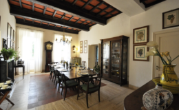 Elegant dining room featuring a long wooden table set for a meal, surrounded by dark wooden chairs. The room boasts high ceilings with exposed beams, large windows allowing natural light, and decorative artwork on the walls. A vintage cabinet and decorative vase add charm to the space.