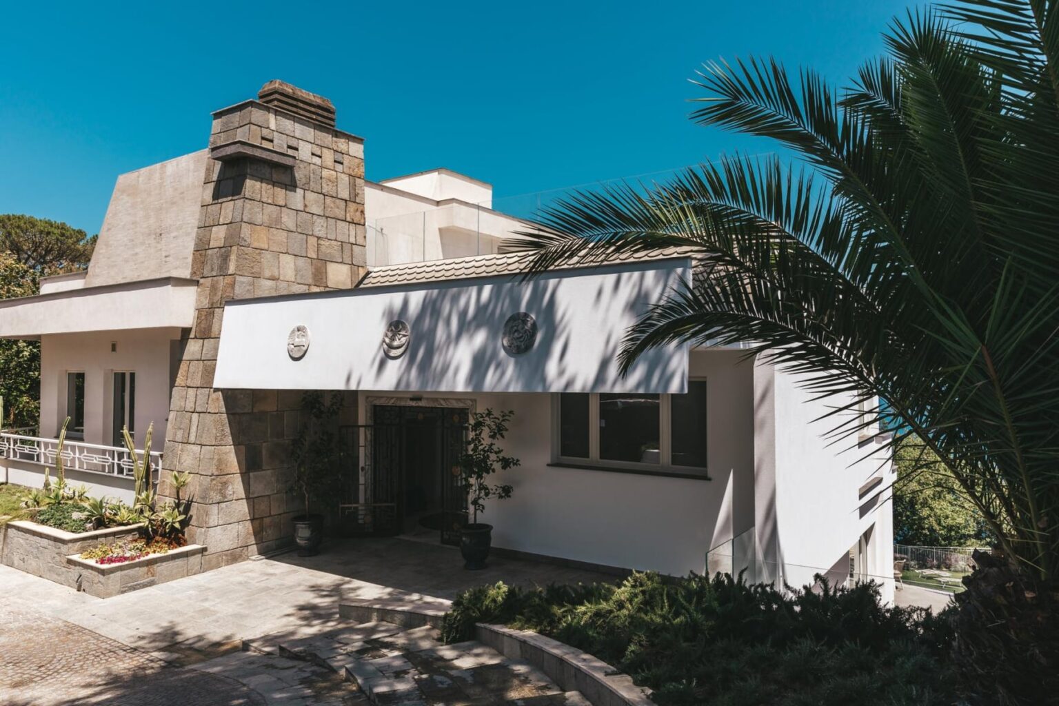 Modern house with a stone chimney, white exterior, and tropical landscaping under a clear blue sky. | Only on SopranoVillas