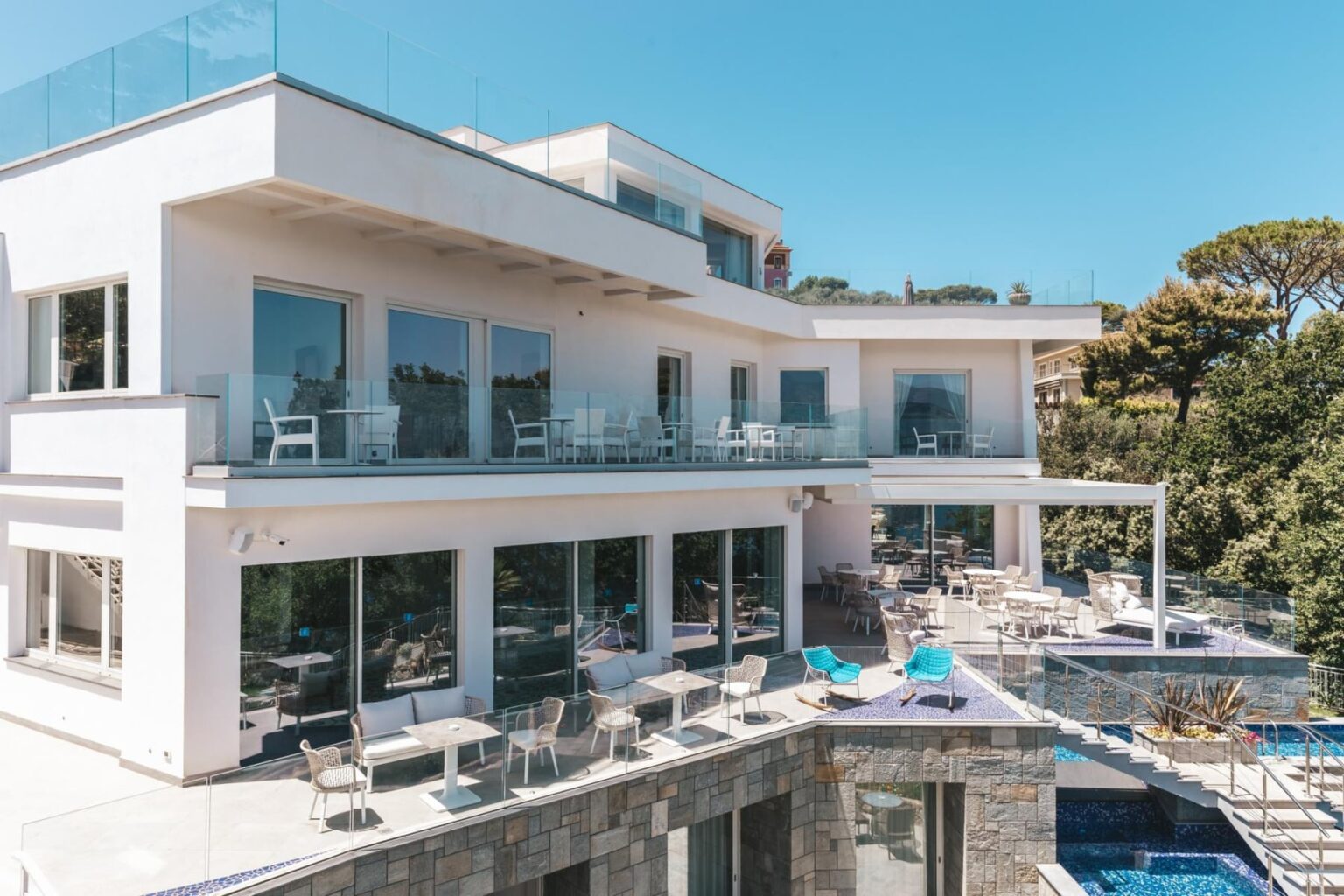 Modern luxury villa with expansive glass windows, outdoor seating areas, and a swimming pool, surrounded by lush greenery and a clear blue sky. | Only on SopranoVillas