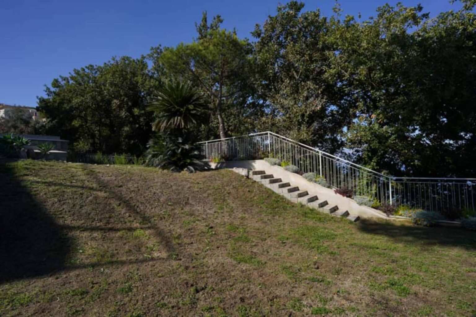 A landscaped garden featuring a grassy area and a set of concrete stairs leading up to a railing, surrounded by lush greenery and trees under a clear blue sky. | Only on SopranoVillas