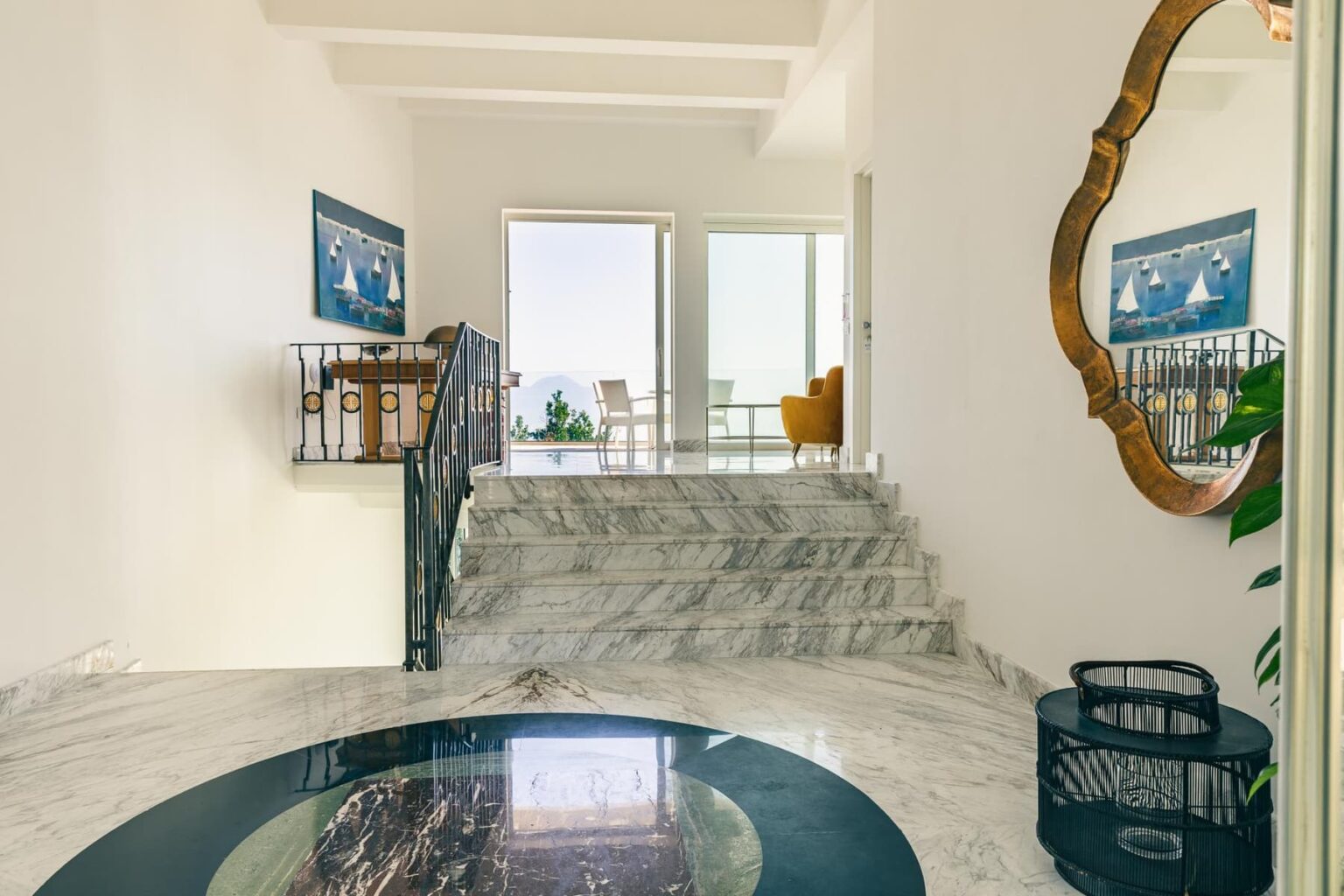 Bright and spacious entryway featuring marble flooring, a decorative round mirror, and a staircase leading to an upper level. Large windows provide natural light and views, complemented by stylish artwork on the walls. | Only on SopranoVillas