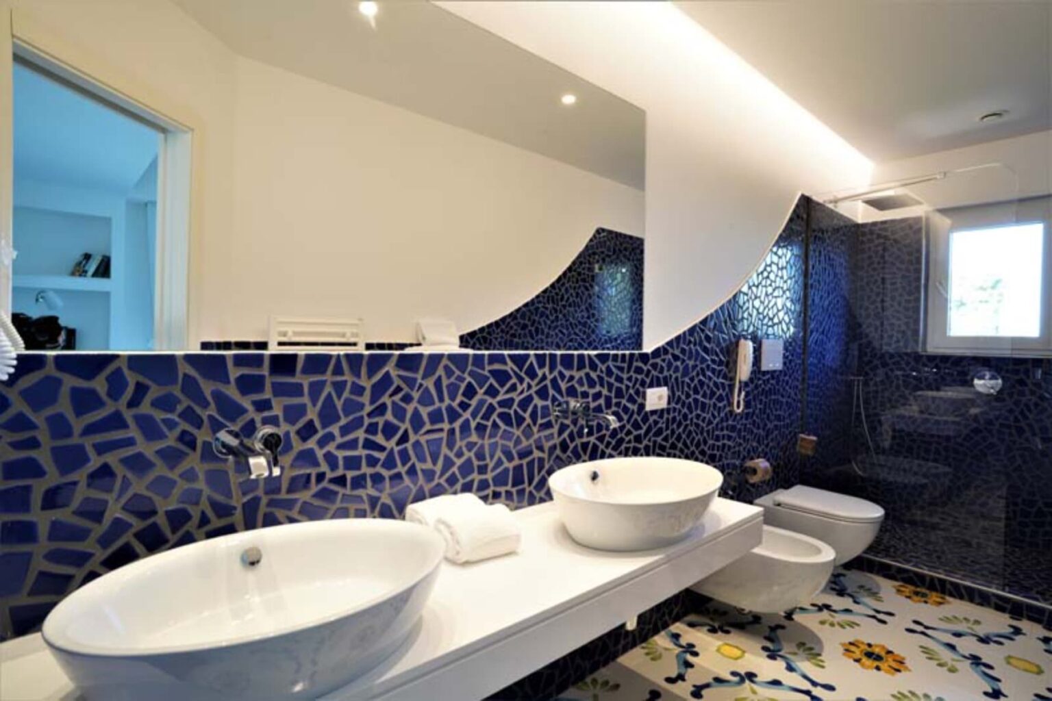Modern bathroom featuring a stylish design with blue mosaic wall tiles, two white vessel sinks on a sleek countertop, and contemporary fixtures. Natural light enters through a window, enhancing the spacious feel. | Only on SopranoVillas