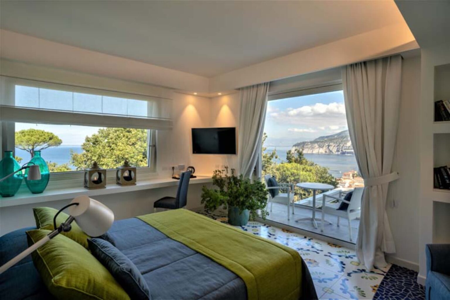 Modern bedroom with a stunning ocean view, featuring a blue and green color scheme, a desk, and decorative plants. Large windows allow natural light to fill the space, enhancing the contemporary decor and inviting atmosphere. | Only on SopranoVillas