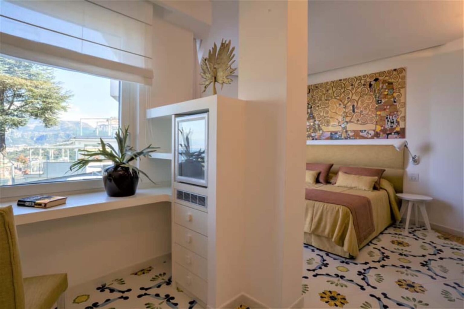 Cozy hotel room featuring a comfortable bed with decorative bedding, a stylish desk area, and large windows offering natural light and a scenic view, complemented by a patterned floor. | Only on SopranoVillas