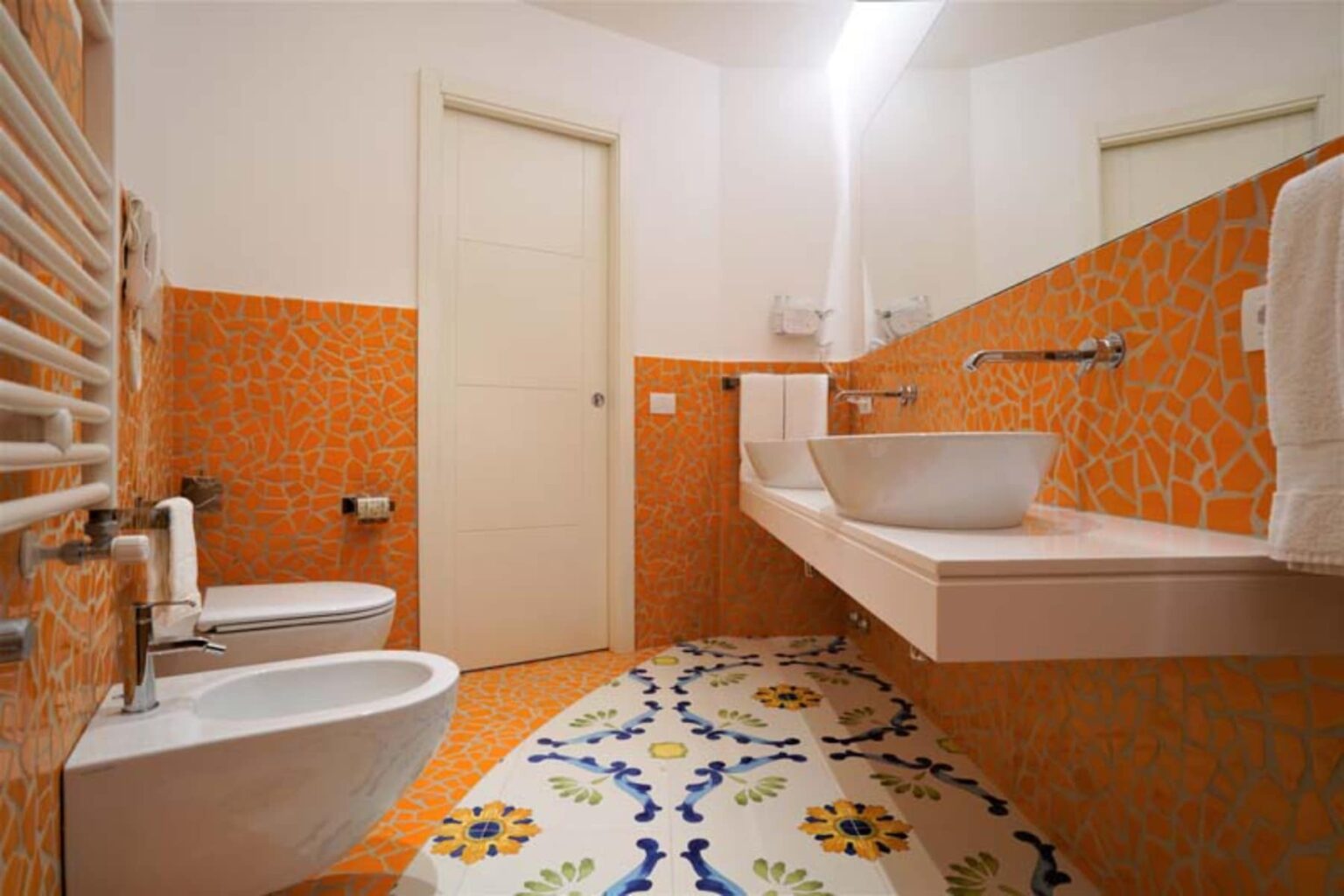 Modern bathroom featuring vibrant orange tiled walls, a sleek white sink with a minimalist design, a bidet, and a decorative floor with colorful patterns. The space is well-lit and has a contemporary feel. | Only on SopranoVillas