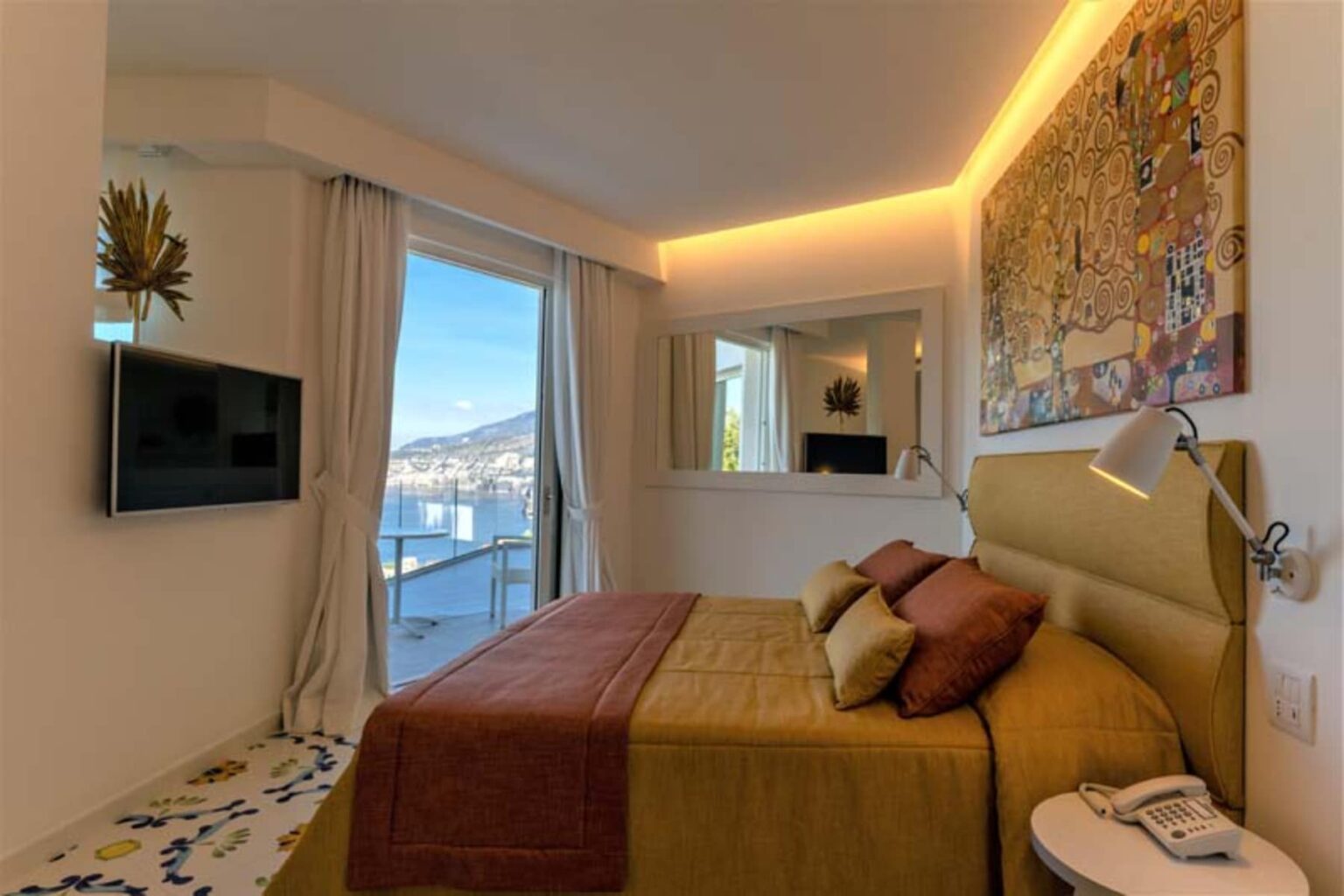 Modern hotel bedroom featuring a queen-sized bed with decorative pillows, a large window with scenic views, a flat-screen TV mounted on the wall, and contemporary decor including a colorful artwork above the bed. The room is well-lit with ambient lighting and stylish furnishings. | Only on SopranoVillas