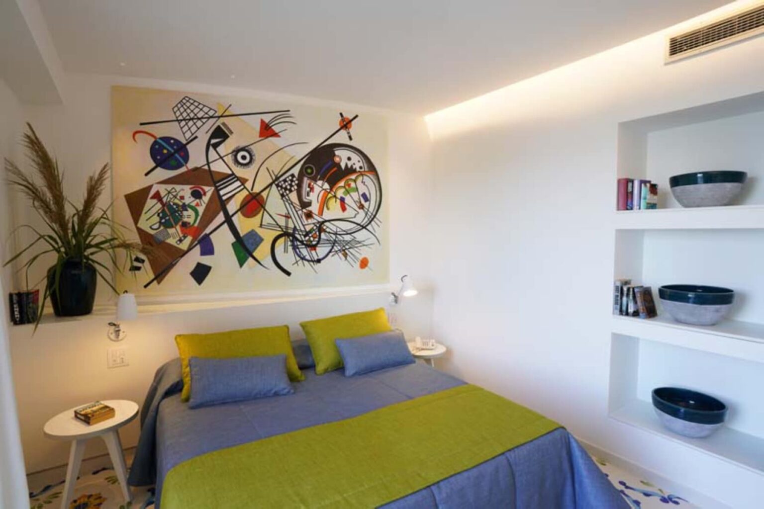 Modern bedroom featuring a colorful abstract wall mural, a double bed with green and blue bedding, stylish nightstands with lamps, and minimalist shelving displaying decorative bowls and books. | Only on SopranoVillas
