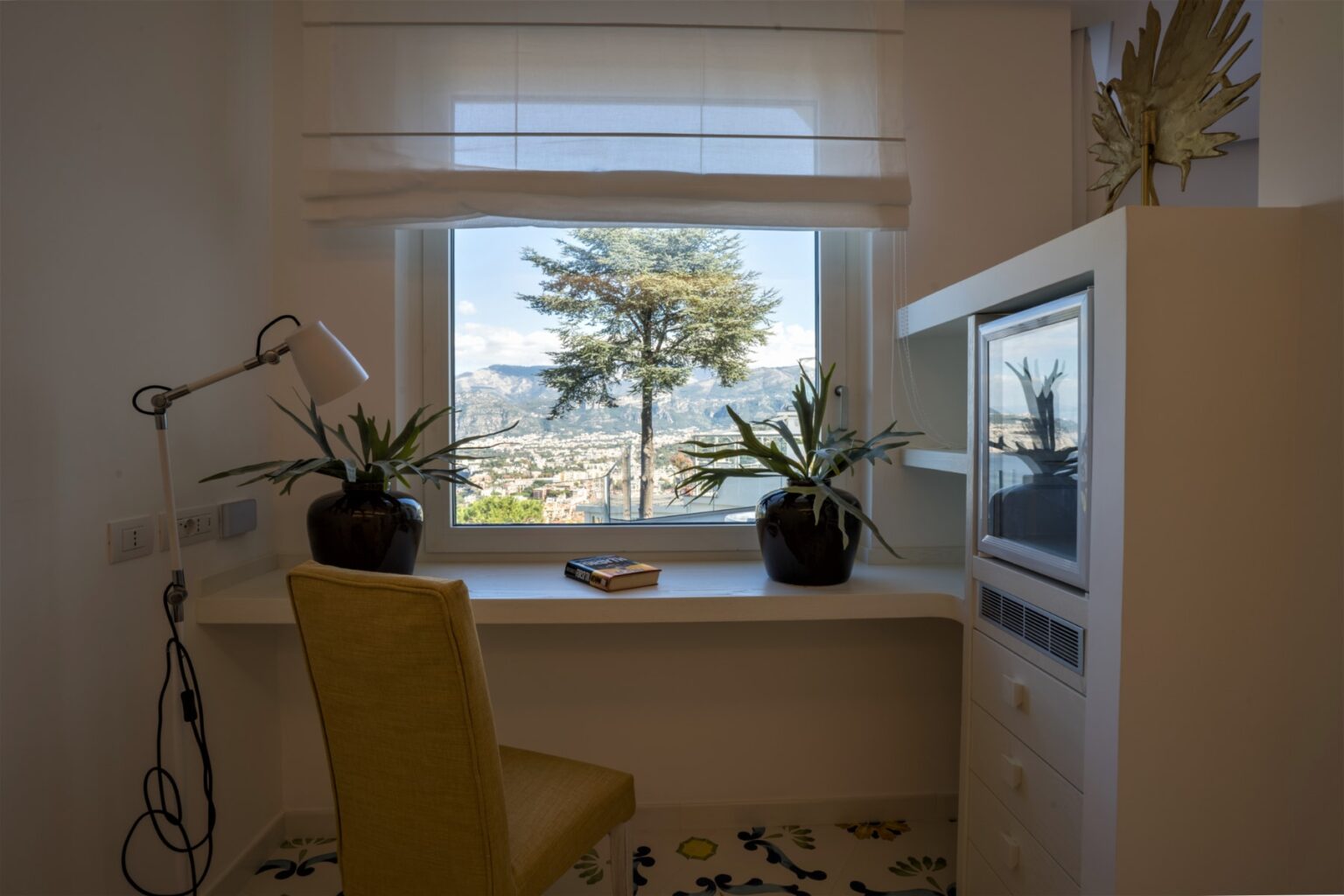 Cozy workspace with a yellow chair, window view of trees and mountains, potted plants on a desk, and a television in a bright room. Ideal for remote work or study. | Only on SopranoVillas