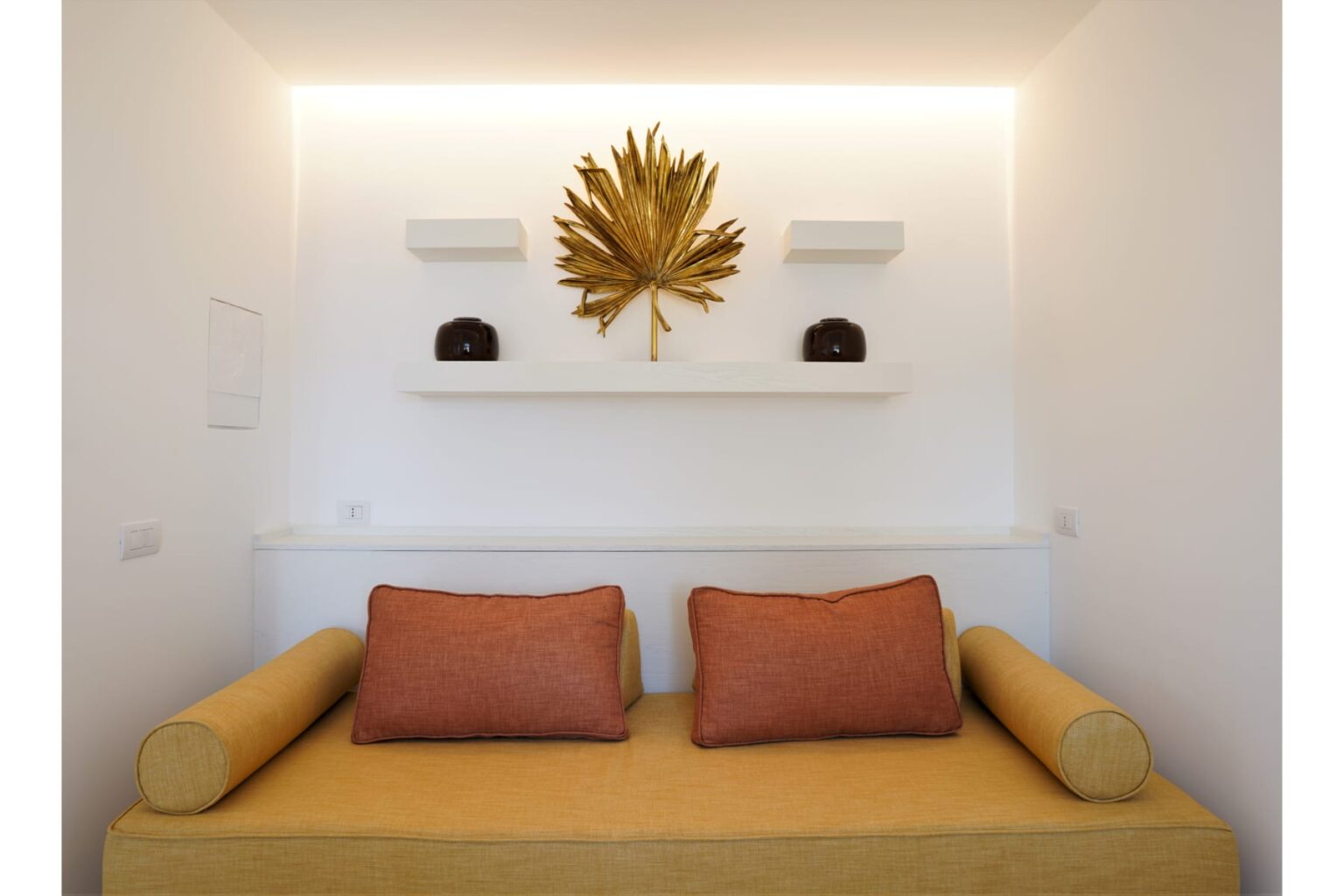 Cozy living space featuring a yellow sofa with orange cushions, adorned with a decorative gold leaf wall art and minimalist white shelves. | Only on SopranoVillas