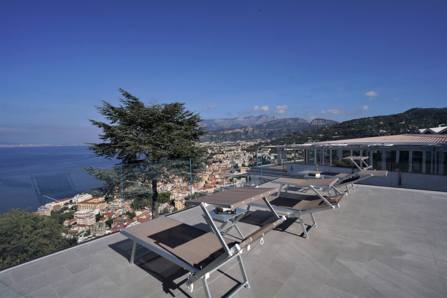 Scenic view from a terrace overlooking a coastal town, featuring lounge chairs and a tree, with mountains and blue sky in the background. | Only on SopranoVillas