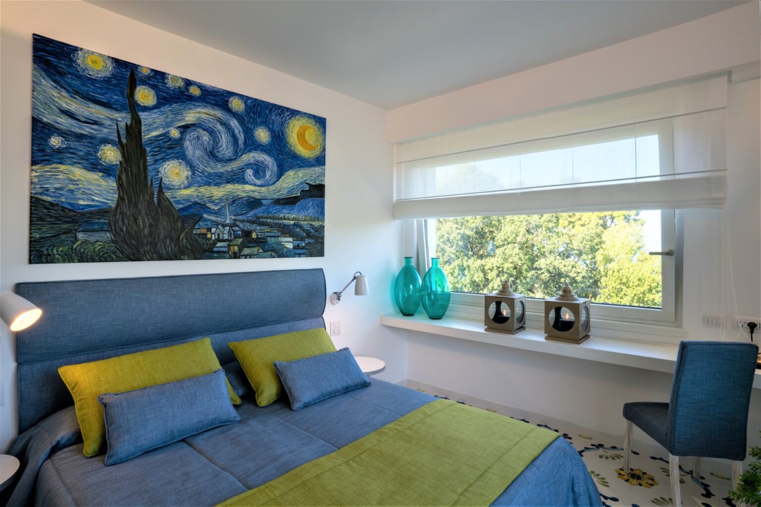 Modern bedroom featuring a blue and green color scheme, with a large reproduction of Van Gogh's "Starry Night" on the wall. The bed is adorned with matching pillows, and natural light streams through a window with sheer blinds, illuminating decorative glass vases and a small lantern on the windowsill. | Only on SopranoVillas