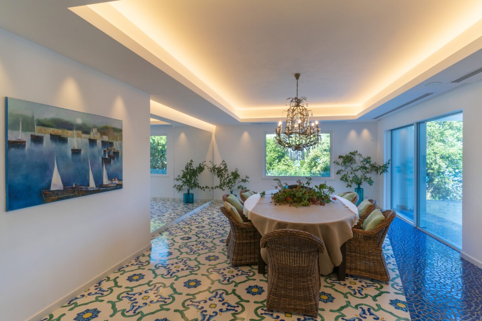 Bright and airy dining room featuring a round table surrounded by wicker chairs, a chandelier, and large windows with greenery visible outside. The floor showcases intricate patterned tiles, complemented by a coastal-themed painting on the wall. | Only on SopranoVillas