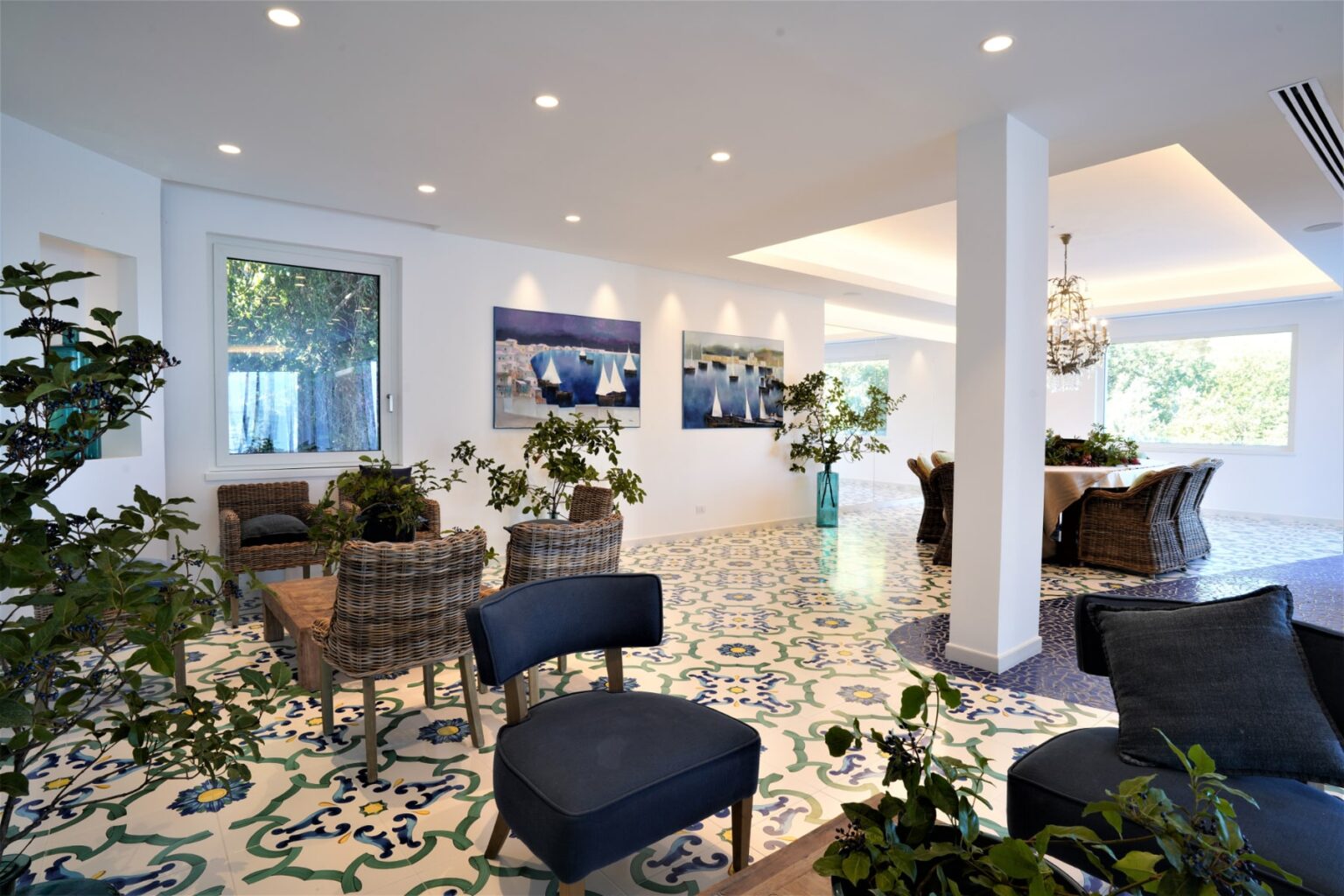 Modern interior design featuring a spacious lounge area with stylish seating, decorative plants, and colorful tiled flooring. Bright natural light illuminates the room, complemented by contemporary artwork on the walls and elegant lighting fixtures. | Only on SopranoVillas