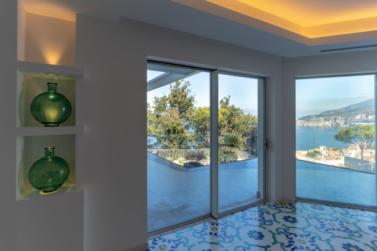 Bright and airy interior featuring green glass vases on a white shelf, large sliding glass doors offering a stunning view of a coastal landscape, and colorful patterned tiles on the floor. Perfect for showcasing modern home decor and scenic views. | Only on SopranoVillas