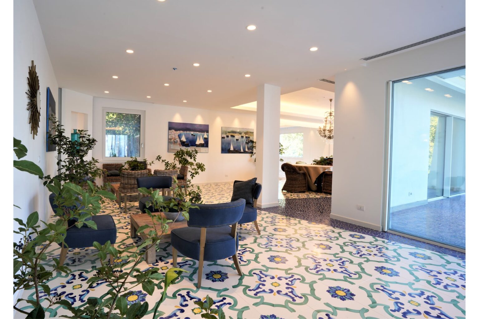 Modern living room with colorful patterned tile flooring, featuring comfortable blue chairs, indoor plants, and large windows that allow natural light to fill the space. | Only on SopranoVillas