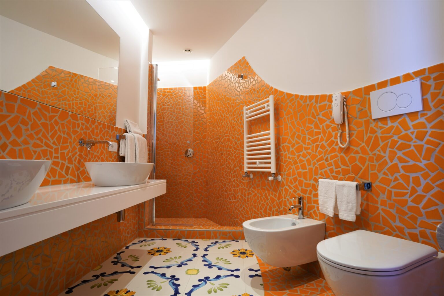 Modern bathroom featuring orange mosaic tiles, a stylish floating sink, a bidet, and a toilet. The space is well-lit with contemporary fixtures and decorative floor tiles. | Only on SopranoVillas