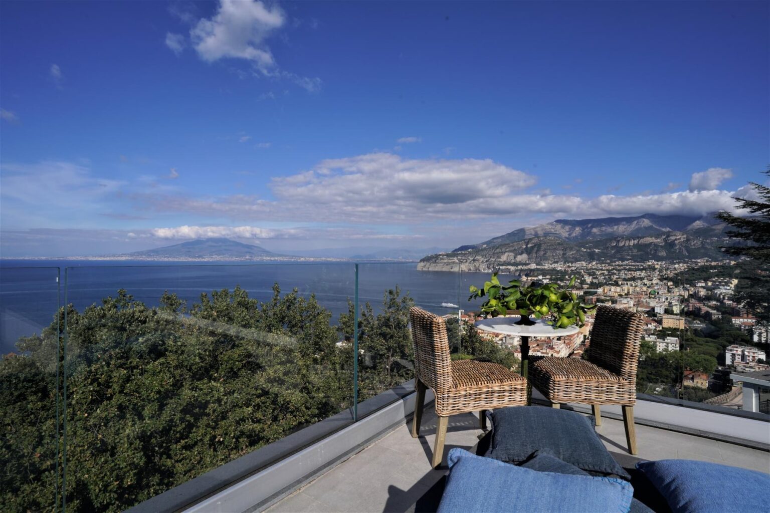 Scenic view from a terrace overlooking the coastline, featuring two wicker chairs and a small table with a plant, framed by a clear blue sky and distant mountains. | Only on SopranoVillas