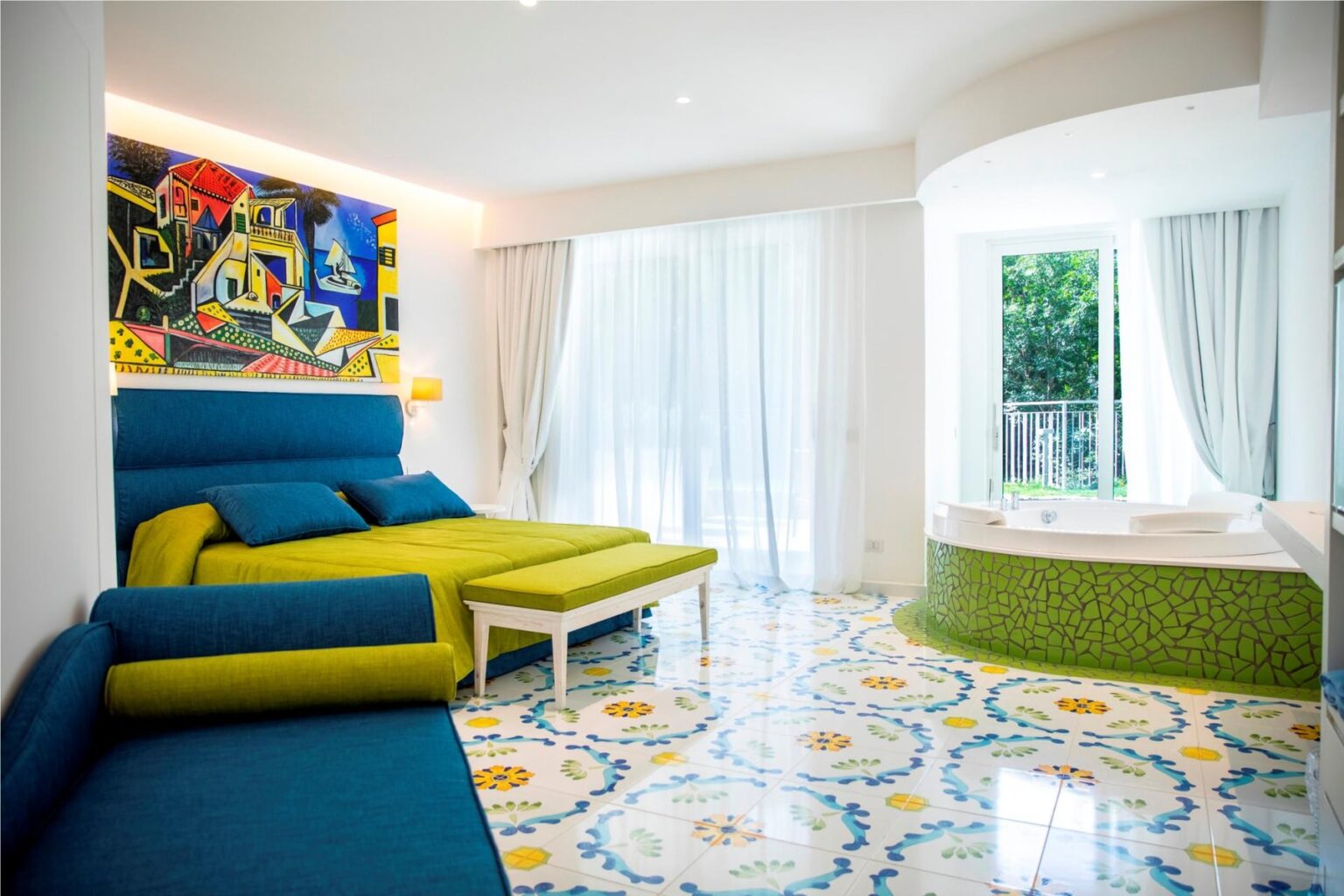 Bright and modern hotel room featuring a blue and green sofa, a white table, and a unique green jacuzzi. Large windows with sheer curtains allow natural light, complemented by colorful tile flooring and vibrant artwork on the wall. | Only on SopranoVillas