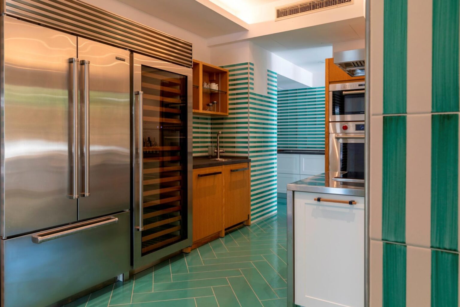 Modern kitchen featuring stainless steel appliances, including a large refrigerator and wine cooler, complemented by wooden cabinetry and vibrant teal tile accents. The layout showcases a sleek design with ample storage and stylish finishes. | Only on SopranoVillas