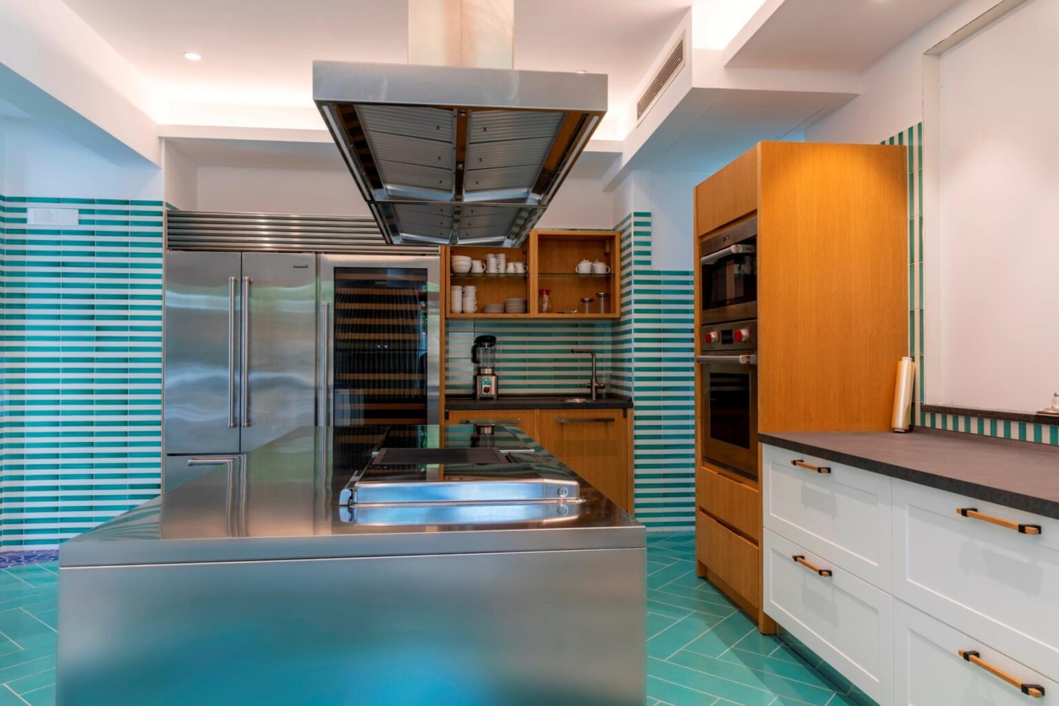 Modern kitchen with stainless steel appliances, a central island with a cooktop, and turquoise tiled flooring. Features wooden cabinetry and stylish backsplash, creating a contemporary and inviting cooking space. | Only on SopranoVillas