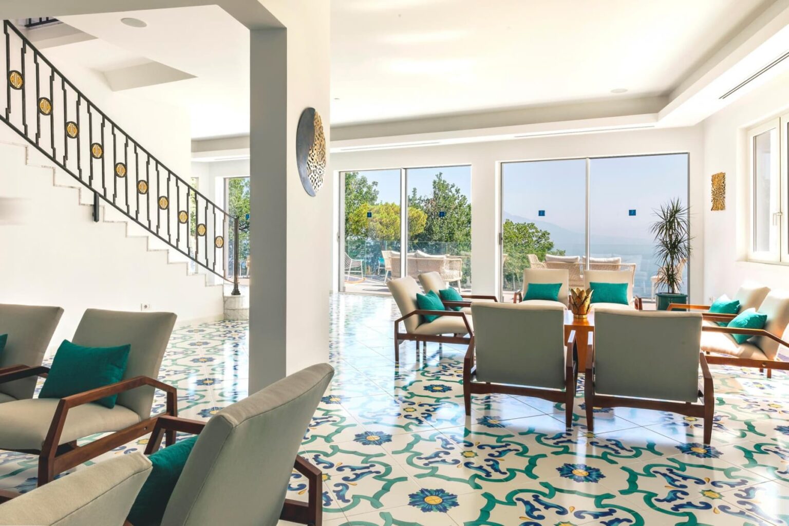Bright and airy interior of a modern living room featuring elegant furniture, vibrant patterned floor tiles, large windows with scenic views, and a stylish staircase, perfect for relaxation and social gatherings. | Only on SopranoVillas