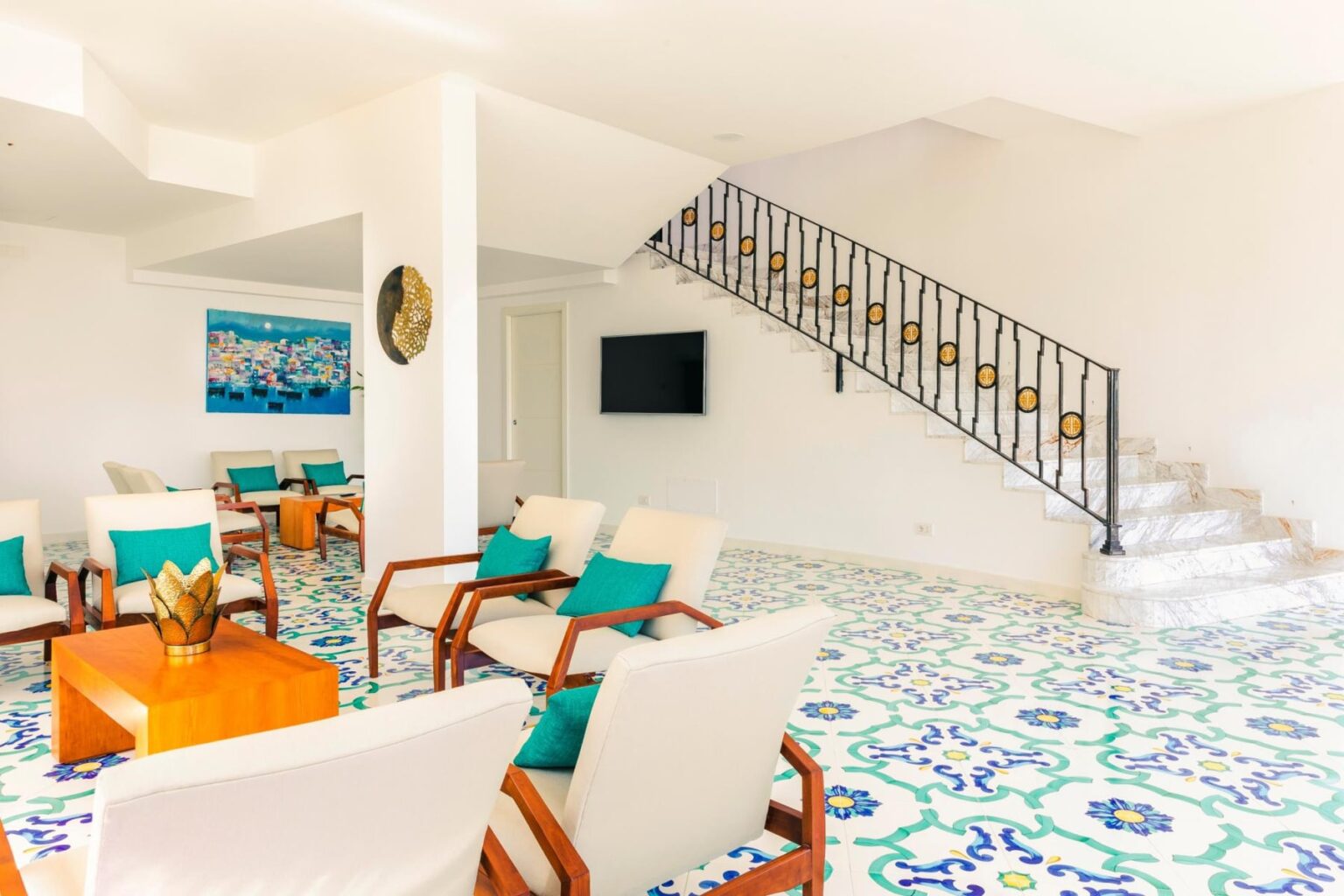 Modern living room with colorful tiled flooring, featuring white seating with turquoise cushions, a wooden coffee table, and a staircase leading to the upper level. A wall-mounted TV and artwork add to the contemporary decor. | Only on SopranoVillas