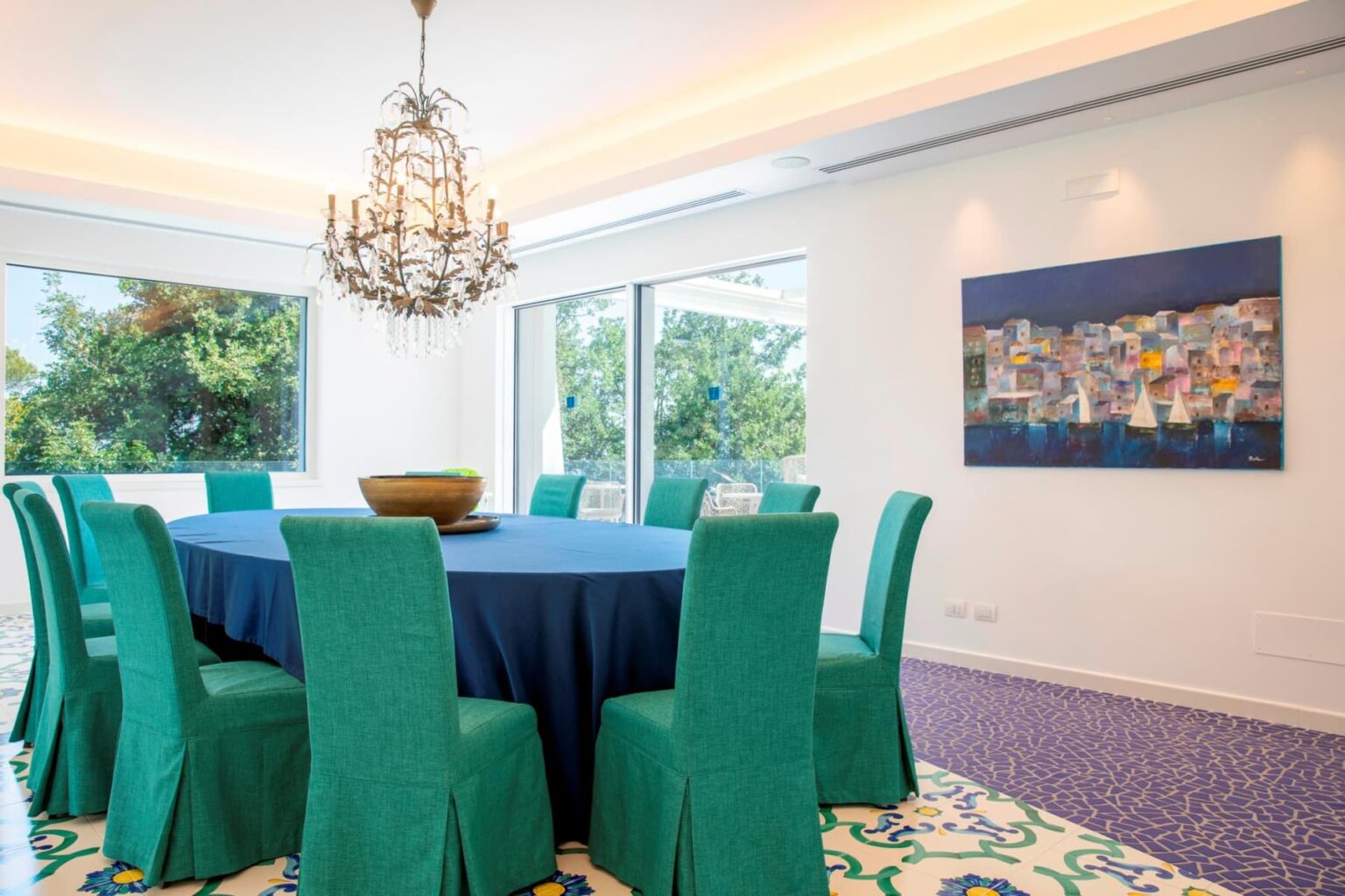 Modern dining room featuring a large round table covered with a dark blue tablecloth, surrounded by vibrant green chairs. A luxurious chandelier hangs from the ceiling, and large windows provide natural light and a view of greenery outside. A colorful painting adorns the wall, enhancing the room's contemporary design. The floor showcases intricate tile patterns, adding to the aesthetic appeal. | Only on SopranoVillas