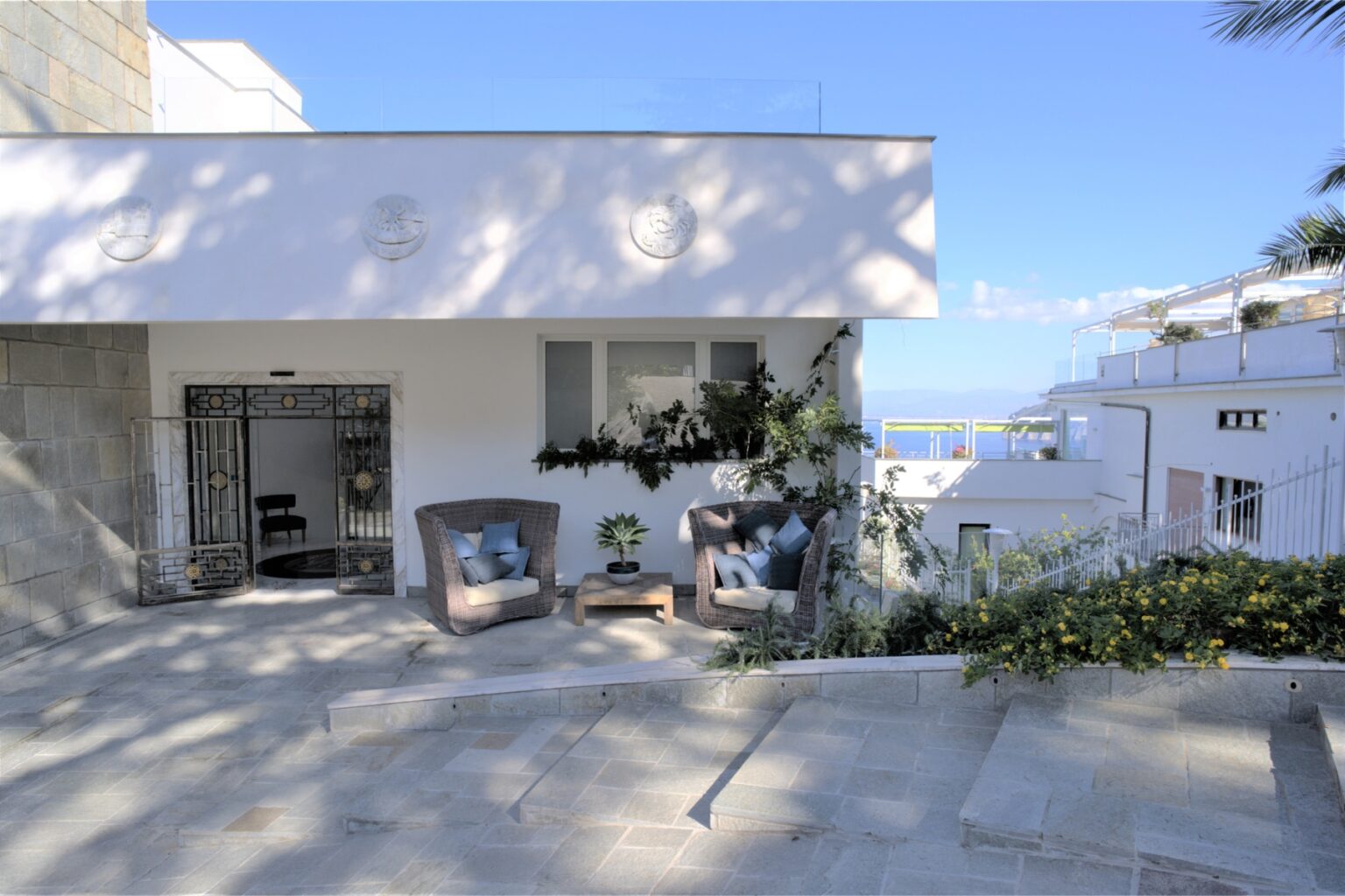 Modern coastal home exterior with a patio featuring comfortable seating and lush greenery, set against a clear blue sky and ocean view. Ideal for outdoor relaxation and entertaining. | Only on SopranoVillas