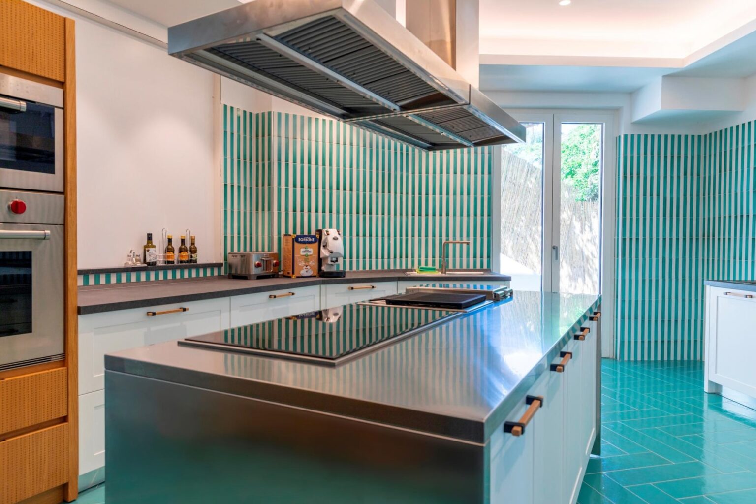 Modern kitchen featuring stainless steel appliances, a large central island with a built-in cooktop, and vibrant green tile accents on the walls and floor. Natural light floods in through large windows, enhancing the contemporary design. | Only on SopranoVillas
