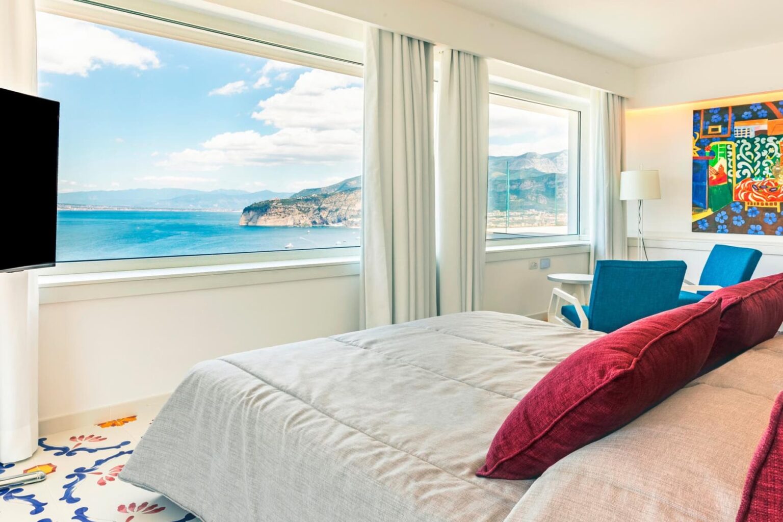 Modern hotel room with a stunning ocean view, featuring a comfortable bed with red pillows, stylish blue chairs, and large windows allowing natural light. | Only on SopranoVillas