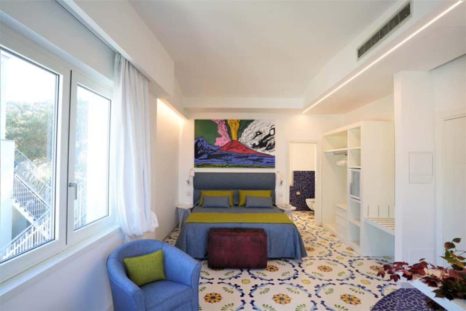 Modern hotel room featuring a vibrant color scheme, a large bed with decorative pillows, artistic wall decor, and a cozy seating area with a blue chair. Natural light floods the space through large windows, complementing the stylish tiled floor and minimalist design elements. | Only on SopranoVillas