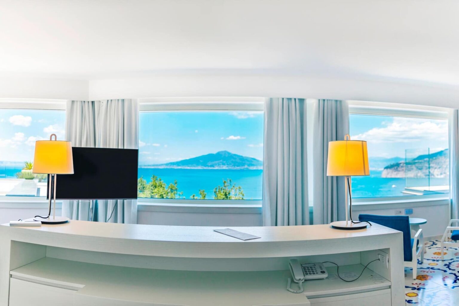 Modern hotel room with panoramic views of the ocean and mountains, featuring a sleek desk, flat-screen TV, and stylish yellow lamps. | Only on SopranoVillas
